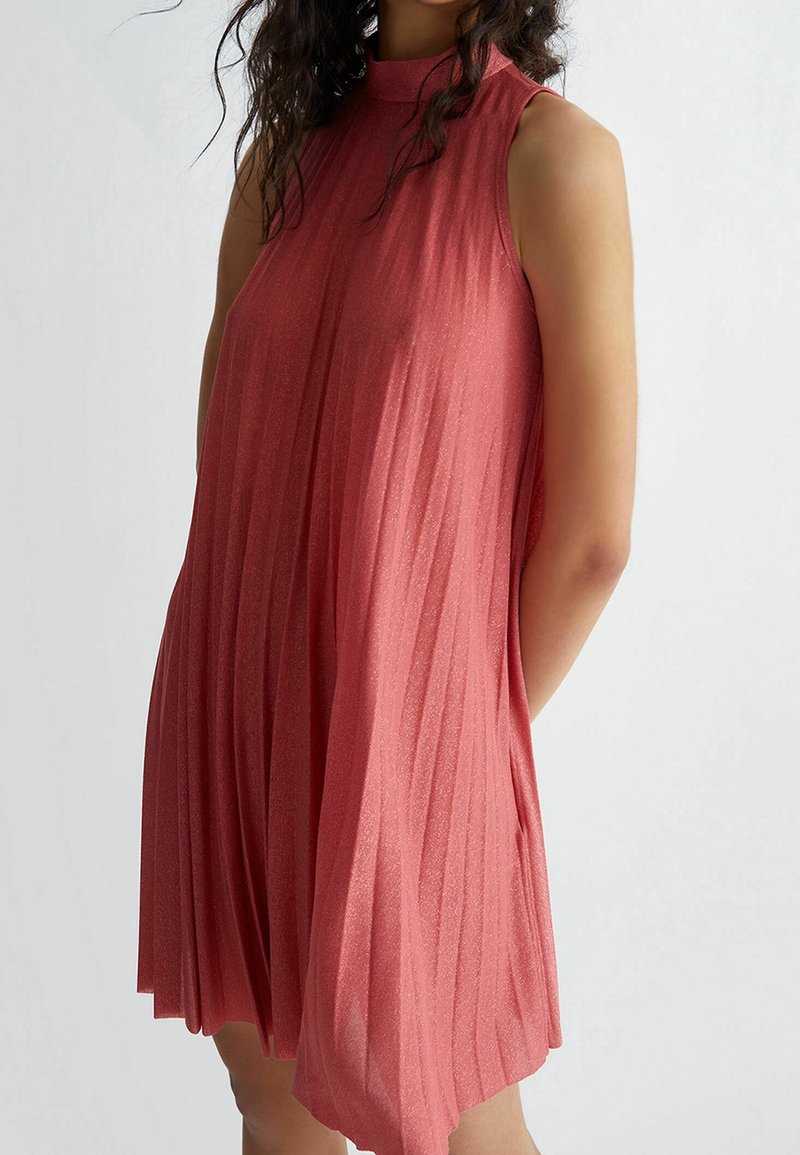 Donna che indossa un vestito senza maniche, a collo alto, plissettato e lucido di colore rosa, in piedi contro uno sfondo chiaro e uniforme.