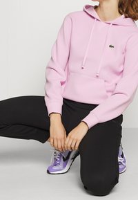 Sudadera rosa hecha de tela suave con un bolsillo tipo canguro y cordones. Un pequeño logo verde está en el pecho. Se lleva con pantalones negros y zapatos morados.