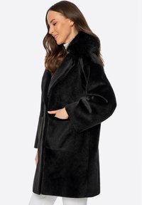 Abrigo de piel sintética negra con un amplio cuello, mangas largas, dos bolsillos frontales y una textura suave, que llega por encima de la rodilla.