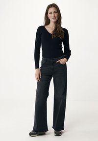 Top lungo a maniche ribatte con scollo a V di colore nero, abbinato a jeans grigi scuri a gamba larga. Il modello sta in piedi con una mano in tasca, indossando scarpe da ginnastica neutre.