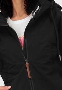 Donna che indossa una giacca nera parzialmente zipata con cappuccio regolabile, che mostra una maglietta bianca con un design floreale rosa e arancione.