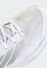 Białe sportowe buty z teksturowaną siateczką, srebrnymi akcentami i jasnymi sznurowadłami. Posiadają formowaną podeszwę oraz wyraźne wzory po bokach.