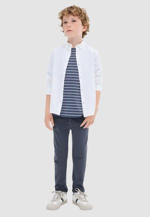 Witte overhemd met knopen over een marineblauw en wit gestreept t-shirt, gecombineerd met donkere broek en witte sneakers met grijze en marineblauwe accenten.