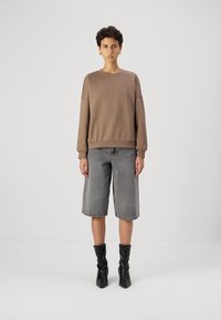 Sudadera marrón con hombros caídos, combinada con pantalones cortos de mezclilla gris. El modelo lleva botas negras hasta el tobillo con un acabado texturizado. Fondo minimalista.