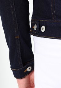 Närbild av ärmen och midjan på en mörk denimjacka med metallknappar, som bärs över vita byxor.