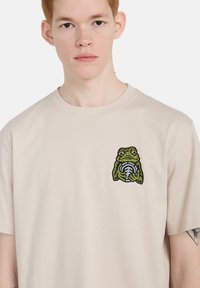 Μπεζ βαμβακερό μπλουζάκι με γραφικό πράσινου βατράχου που κρατάει ένα στρογγυλό αντικείμενο, στρογγυλή λαιμόκοψη, κοντά μανίκια και άνετη εφαρμογή.