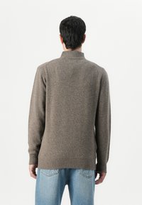 Man in een bruin geribbelde hoge hals trui en lichtblauwe spijkerbroek, met zijn rug naar de camera gekeerd tegen een witte achtergrond.