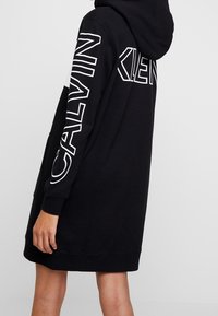 Černé šaty-svetr s kapucí, s bílým textem "CALVIN KLEIN" na rukávech a měkkou, texturovanou tkaninou. Dlouhý, volný střih.