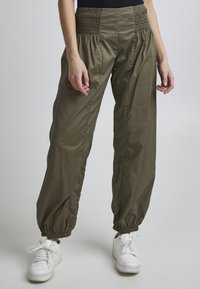 Pantalons vert olive, amples, avec une taille froncée et des poignets élastiques, fabriqués dans un matériau léger. Portés avec des baskets blanches.