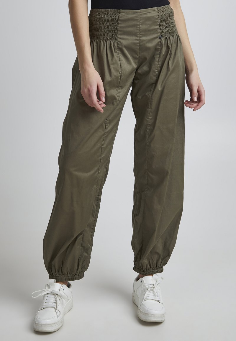 Pantaloni verde oliva, ampi, con vita smock e polsini elasticizzati, realizzati in materiale leggero. Indossati con sneakers bianche.