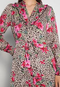 Abito con motivo floreale che presenta rose rosa e foglie verdi su uno sfondo a stampa leopardo beige e nero. Collo arricciato, bottoni e vita legata.