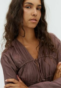 Brown, textured blouse with gathered detail near the neckline. Features a V-neck and ties at the front. Long sleeves and relaxed fit.
