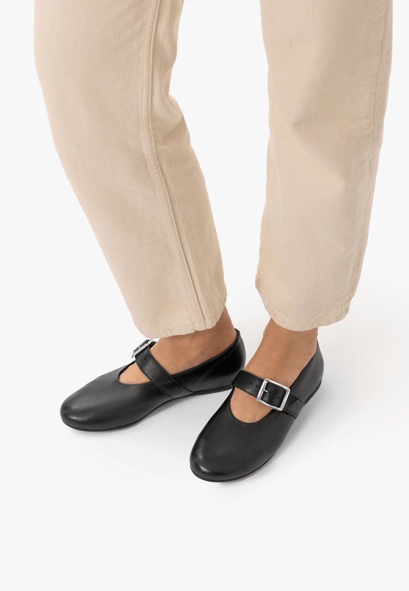 Pieds chaussés de chaussures style Mary Jane noires avec boucles argentées, associés à un pantalon beige clair court sur fond blanc.