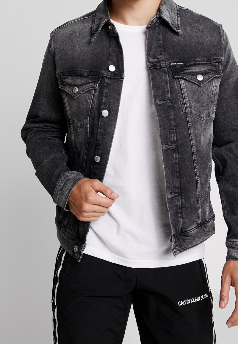 Mörkgrå denimjacka med knäppning fram, två bröstfickor och sliten detalj, bärs över en vit t-shirt och svarta byxor.