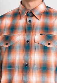 Chemise à carreaux orange, bleue et blanche. Elle comporte un col boutonné, deux poches poitrine avec rabats, et des boutons contrastants. Logo G-Star présent.