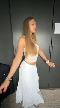 Jeune femme aux longs cheveux blonds portant un débardeur beige et une jupe blanche, debout et étendant les bras dans une pièce avec des armoires sombres.