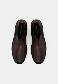Sapatos castanhos de pele, tipo slip-on, com uma superfície lisa, bico arredondado, sola preta e uma marcação dourada na palmilha e lateral.