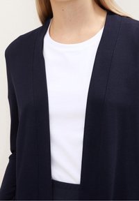 Cardigan aperto blu navy realizzato in tessuto a maglia, con una texture liscia e maniche lunghe, sovrapposto a una maglietta bianca girocollo.