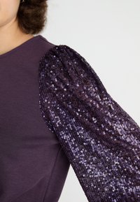 Violet ilgomis rankovėmis palaidinė, pasižyminti prigludusia medžiaga ir dideliu blizgučių pūstinimu, pagamintu iš įvairių violetinės spalvos atspalvių blizgučių.