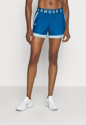 Osoba w niebieskich sportowych szortach Under Armour i białych butach Nike, stojąca na tle prostego jasnego tła.