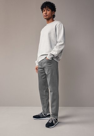 Ein junger Mann trägt ein hellgraues Sweatshirt, graue Hose und schwarze Sneakers und steht mit einer Hand in der Tasche vor neutralem Hintergrund.