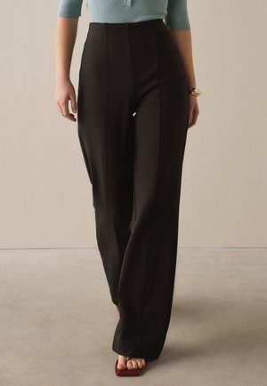 Frau trägt schwarze, hoch taillierte, weite Hose, türkisfarbenes geripptes Oberteil, goldenes Armband und bordeauxrote offene Sandalen, steht drinnen.