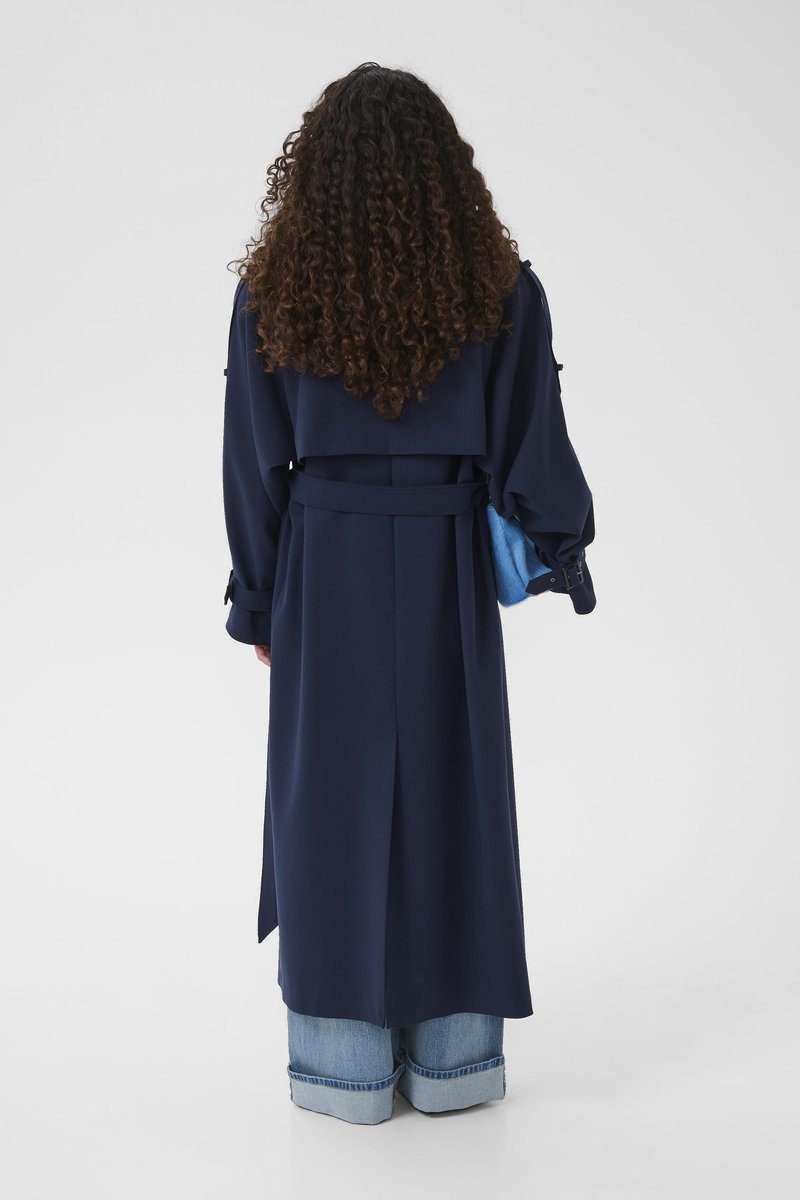 Trench-coat bleu marine avec taille ceinturée, manches larges et détails aux épaules, associé à des pochettes bleu clair et un jean retroussé à revers.