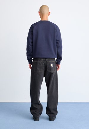 Marineblå hettegenser med raglanermer, kombinert med løst sittende svarte jeans med etikett på baklommen. Enkel design, ingen mønstre.