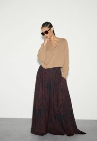 Brązowy sweter dzianinowy z detalami guziczków, zestawiony z ciemno-maruńczową, wzorzystą spódnicą maxi o zwiewnym kroju. Klasyczne czarne okulary przeciwsłoneczne.