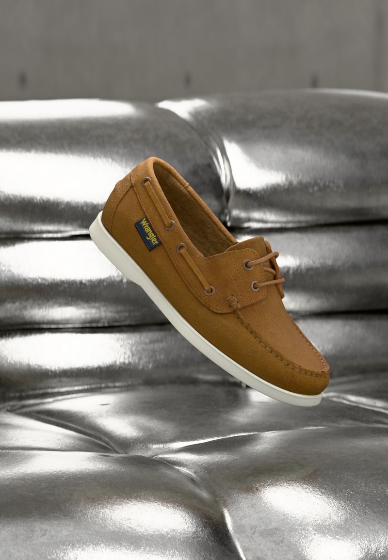 Chaussure style mocassin en daim marron avec semelle blanche et lacets, posée sur un coussin en cuir argenté réfléchissant.