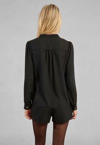 Maglietta nera a maniche lunghe con colletto, design con schiena plissettata, polsini con bottoni e una texture liscia su uno sfondo neutro.