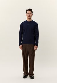 Álló férfi sötétkék hosszú ujjú henley ingben, barna nadrágban és fekete cipőben, egyszerű világos háttér előtt.