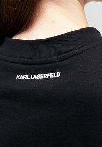 Prenda de algodón negro con cuello acanalado. Presenta el logotipo en blanco "KARL LAGERFELD" en la parte posterior, cerca del escote. Textura suave.