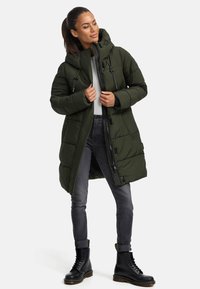 Green puffer coat with a hood, featuring a zip closure and drawstrings. Worn over a light top, paired with gray jeans and black boots.