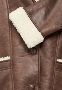 Giacca in ecopelle marrone con colletto e polsini in shearling testurizzato crema, due tasche frontali e bottoni marroni scuri.