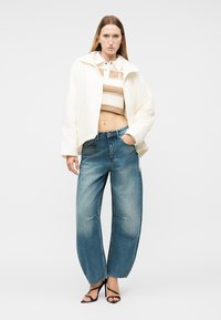 Vrouw draagt een wit cropped jasje over een beige gestreepte cropped polo, losse blauwe spijkerbroek en zwarte sandalen met bandjes, staand tegen een witte achtergrond.