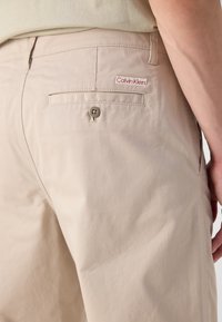 Beige bomullsbukser med en baklomme, med rund knappelukking og et lite rødt Calvin Klein-logo-lapp. Glatt tekstur.