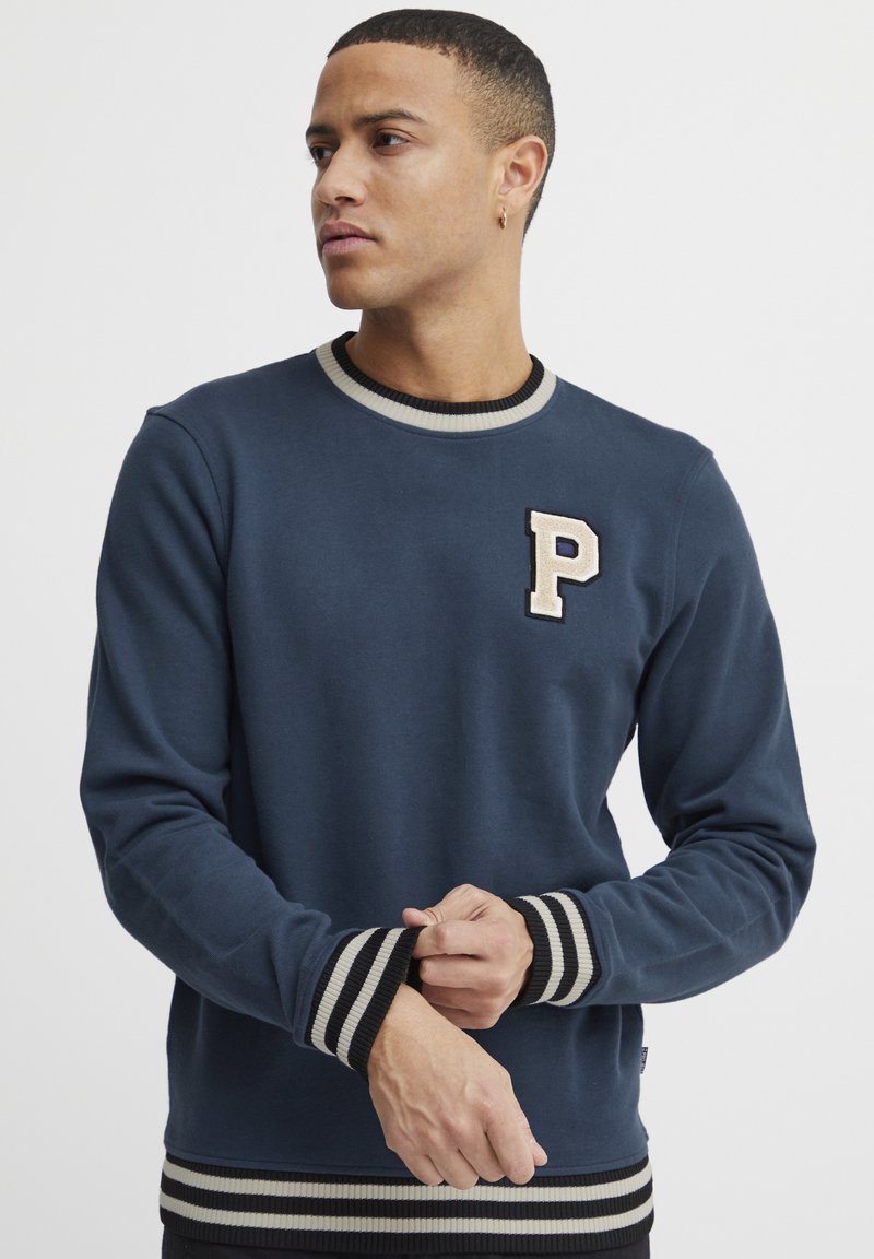 Marine sweatshirt gemaakt van zachte stof, met een crème en zwarte gestreepte geribbelde kraag en manchetten, en een decoratieve "P" patch op de borst.