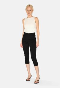 Biely bezrukávový top s jemným vzorom, kombinovaný s čiernymi cropped nohavicami. Model má na sebe čierne žabky. Nevýrazné pozadie.