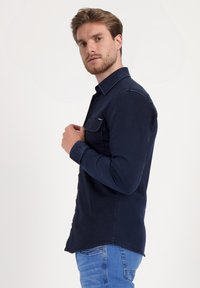 Camisa azul marino de botón con cuello clásico, bolsillo en el pecho y mangas largas, con un dobladillo ligeramente curvado y textura suave.
