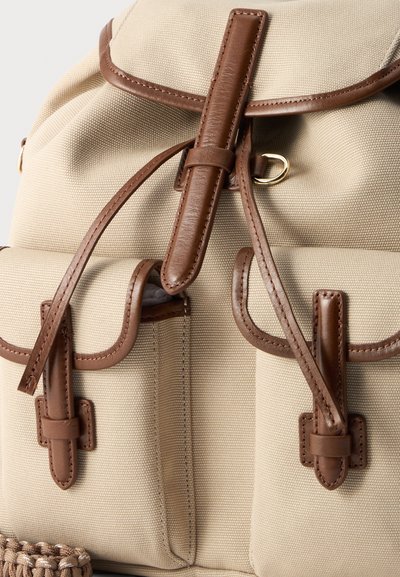 Sac à dos en toile beige avec garniture et sangles en cuir marron, doté de deux poches avant à fermeture par boucle et d'un haut à cordon de serrage.