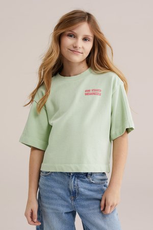 Dziewczyna o długich blond włosach, ubrana w jasnobeżową, oversize'ową koszulkę z napisem "Sun Kissed Memories" oraz luźne niebieskie dżinsy, pozująca na beżowym tle.