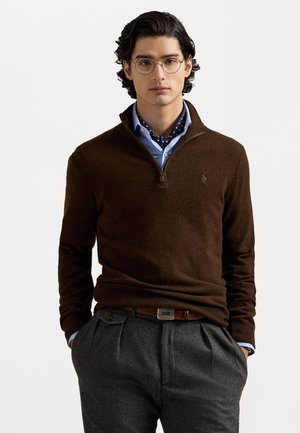 Pull zippé marron en maille avec un ourlet côtelé, superposé à une chemise bleu clair avec une écharpe à pois navy. Pantalons gris à plis.