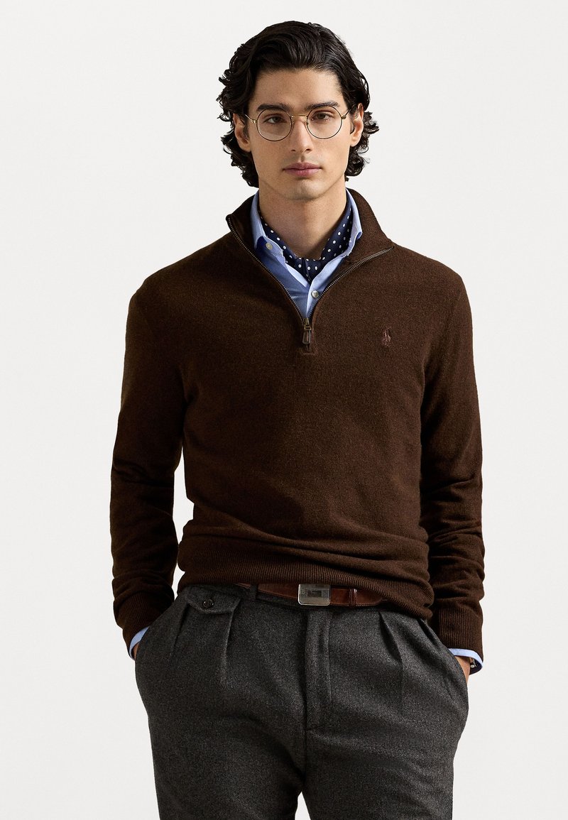 Suéter marrón con cuello de cremallera, de material de punto y con dobladillo de canalé, superpuesto a una camisa azul claro y acompañado de una bufanda con estampado de lunares en navy. Pantalones grises con pliegues.