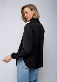 Czarna satynowa bluzka z długim rękawem, o wysokim kołnierzu i lekko luźnym kroju; dżinsy w odcieniu vintage, poddane praniu w pralce.