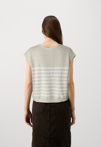 Kobieta z długimi brązowymi włosami, ubrana w beżowy dzianinowy top bez rękawów z białymi poziomymi paskami i ciemnobrązową spódnicą, odwrócona tyłem.