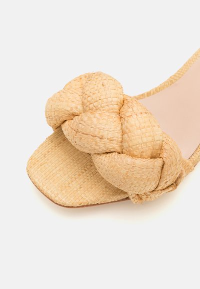 Vævede stråslidesandaler med et stort knyttet design. Sandalen har en flad sål og et tekstureret, naturligt beige materiale.