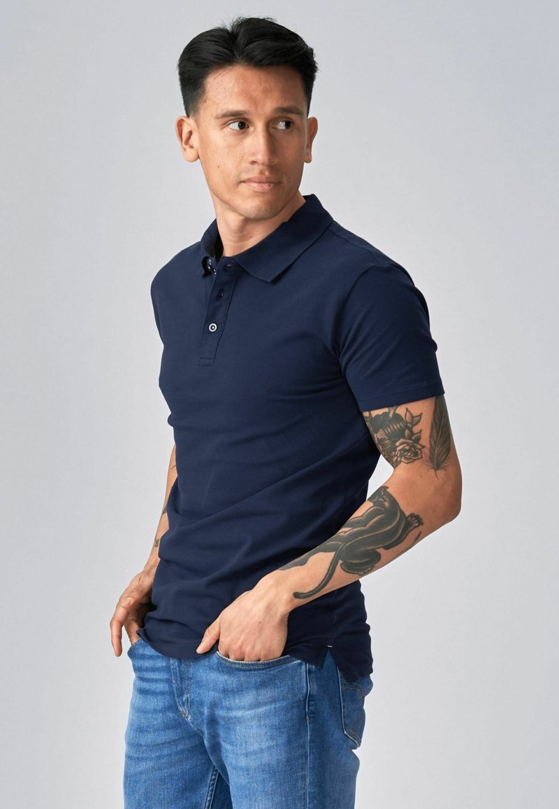 Hombre con cabello oscuro vistiendo una camiseta polo azul marino y jeans azules, mostrando tatuajes de flores y una pantera en sus brazos, posando con las manos en los bolsillos.