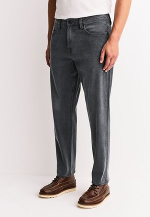 Mand iført mørkegrå jeans med lige ben, hvid skjorte og brune lædersnøresko, stående på en hvid baggrund.