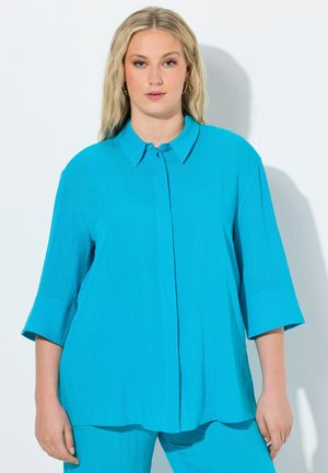 Femme aux longs cheveux blonds portant une chemise turquoise ample à boutons et un pantalon assorti, debout devant un fond blanc.